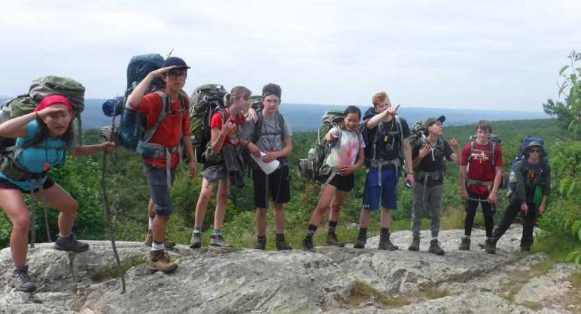 A group of young people wearing backpacks pose for a photo on a rock ledge with a vast green landscape behind them. 